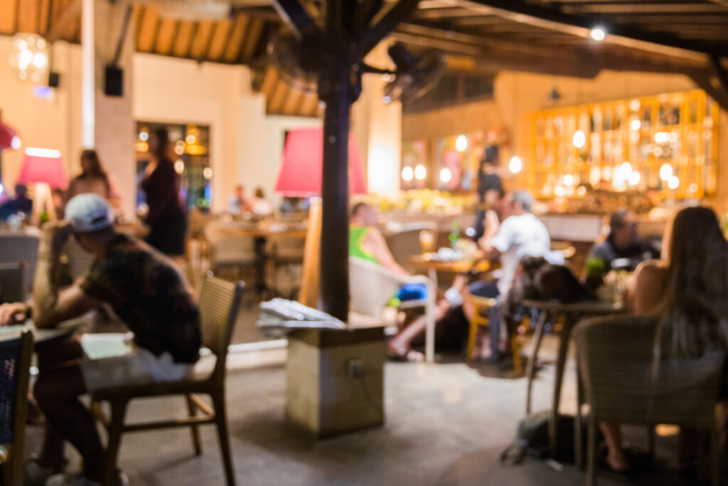 Imagem tem um blur de efeito, mas dá para perceber pessoas sentadas em várias mesas de um bar ou restaurante