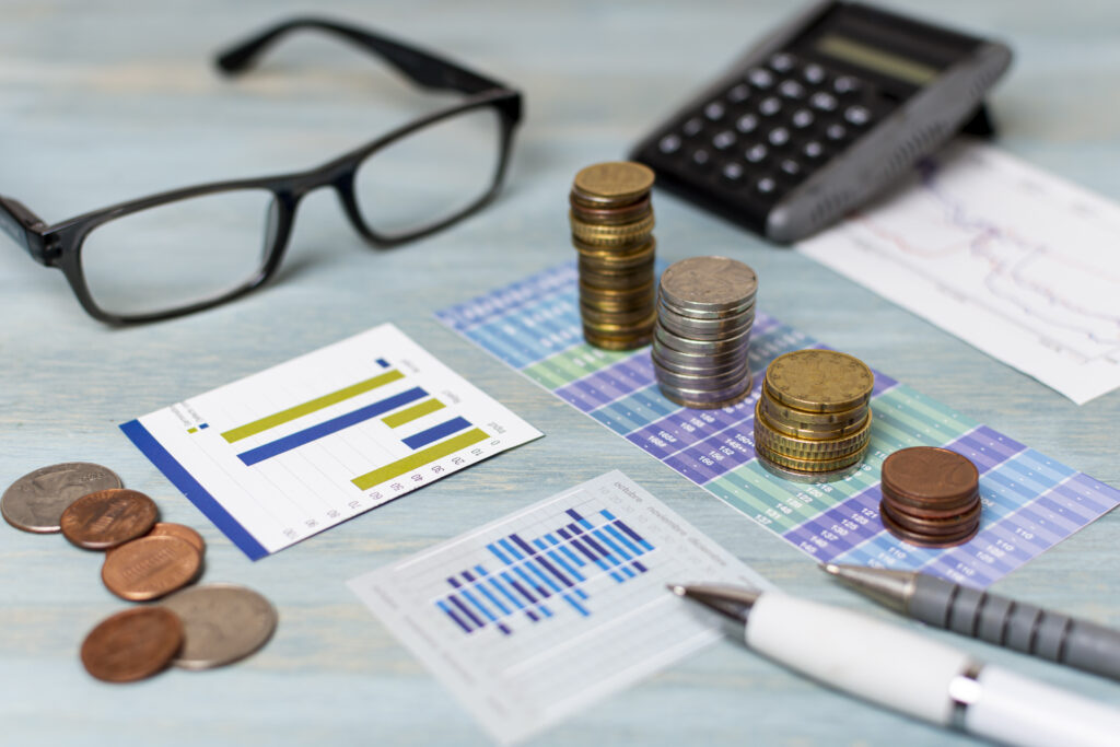 Em uma mesa estão um óculos, moedas, gráficos e calculadora.