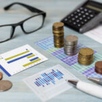 Em uma mesa estão um óculos, moedas, gráficos e calculadora.