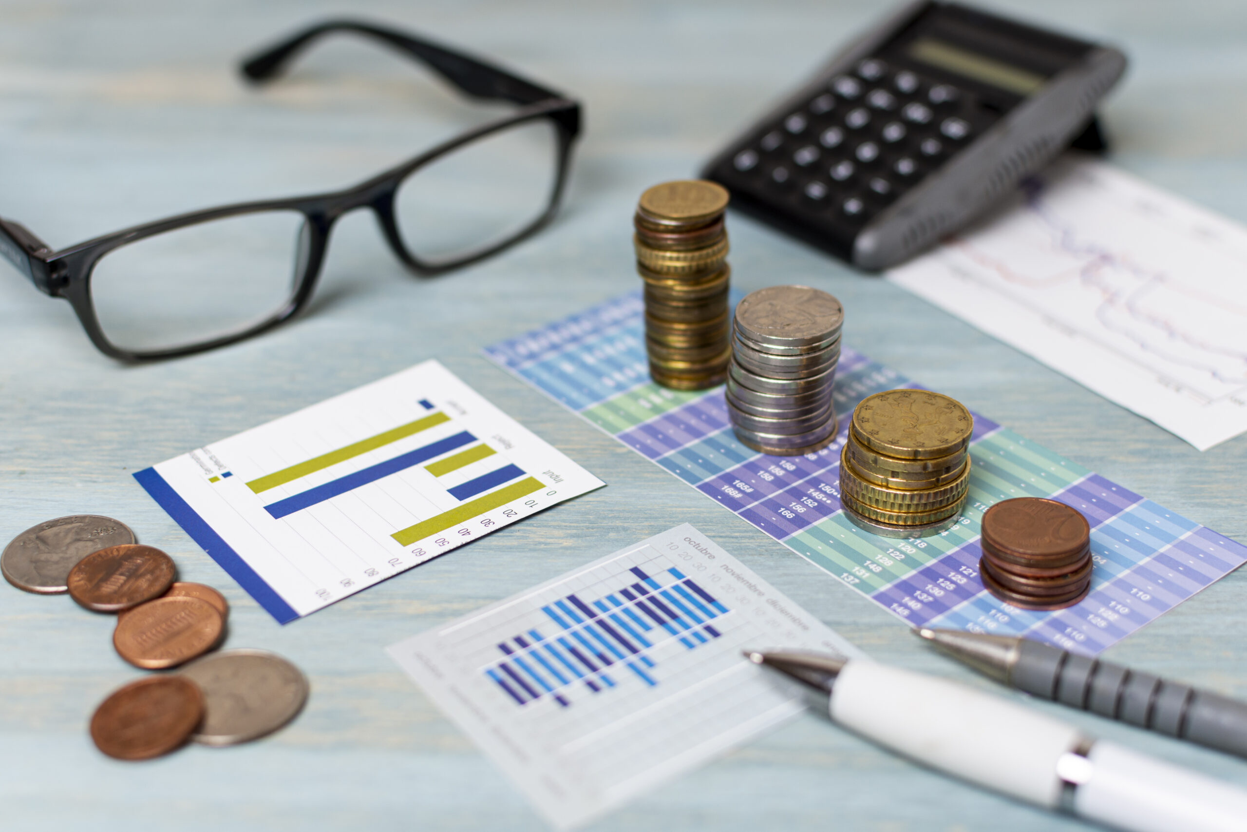 Em uma mesa estão um óculos, moedas, gráficos e calculadora.